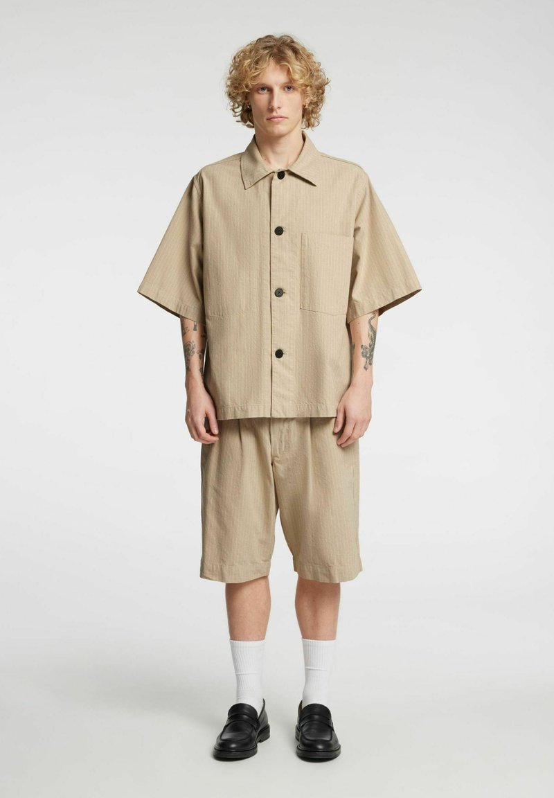 Beige kortermet skjorte med brystlomme og svarte knapper, kombinert med matchende beige shorts, brukt med hvite sokker og svarte loafers.