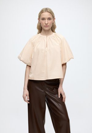 Frau mit glattem blondem Haar trägt eine hellpfirsichfarbene Bluse mit Puffärmeln und dunkelbraune, weit geschnittene Lederhosen, steht vor einem schlichten Hintergrund.