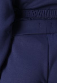 Jūras zila sporta apģērba komplekts izceļas ar mīkstu audumu, elastīgu jostasvietu un maigu tekstūru. Šuves piešķir struktūru dizainam.