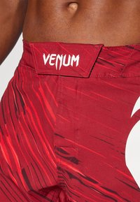 Piros sportnadrág textúrázott mintával, fekete csíkokkal és fehér "Venum" logóval a derékrészen. Tartós anyag, testhezálló szabás.