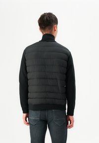 Black puffer vest with horizontal quilted pattern, high collar, and ribbed knit sleeves, worn over a black long-sleeve shirt and dark jeans.