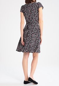 Žena v černých květovaných šatech nad kolena s krátkým rukávem a černých balerínách, stojící na pozadí bílé stěny.