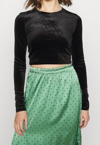 Kvinna iklädd en svart långärmad sammetcroptopp och en hög midja av grön satin med svarta prickar, stående mot en neutral bakgrund.