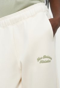 Kremfargede joggebukser laget av mykt materiale, med elastisk midjebånd og brodert grønt "New Balance Athletics"-logo.