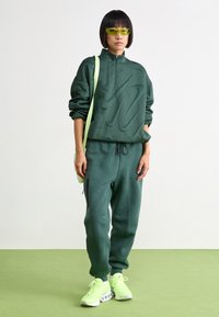 Πράσινη μπλούζα με ζιγκ ζαγκ μοτίβα, συνδυασμένη με αντίστοιχες φόρμες. Τα αξεσουάρ περιλαμβάνουν μια ανοιχτό πράσινη τσάντα χιαστί και νέον πράσινα αθλητικά παπούτσια.