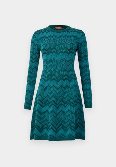 Trikotāžas kleita tirkīza kolorā ar zigzaga rakstu, ar garām piedurknēm un apaļu kakla izgriezumu. Tai ir pieguļošs augšdaļa un izpletusies svārku dizains.