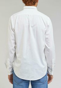 Chemise blanche à manches longues avec col à boutons, ourlet arrondi et poignets boutonnés, en tissu lisse. Portée avec un jean bleu.