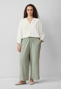 Blouse blanche à manches longues avec décolleté en V, associée à un pantalon léger vert clair ample avec taille élastique et poches latérales. Chaussures plates à motif.