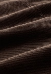 Brown corduroy fabric featuring distinct vertical ridges, smooth texture, and folded layers creating shadows and depth.