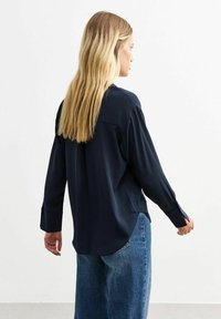 Blusa a maniche lunghe blu navy con vestibilità rilassata, dotata di polsini con bottoni e orlo curvo, realizzata in un tessuto leggero e liscio.