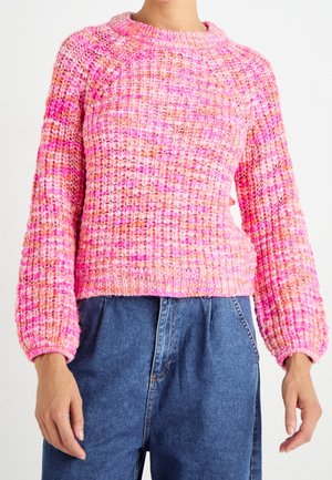 Personne portant un pull en maille épaisse aux tons roses et orange avec des manches longues, associé à un jean en denim bleu taille haute.