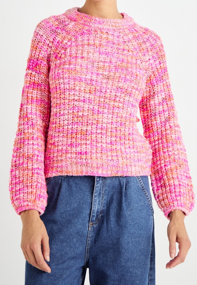 Personne portant un pull en maille épaisse aux tons roses et orange avec des manches longues, associé à un jean en denim bleu taille haute.