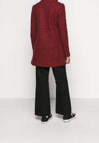 Abrigo de punto rojo, de longitud media hasta el muslo, con un cuello alto. Combina con pantalones negros de pierna ancha y zapatillas negras con suelas blancas.