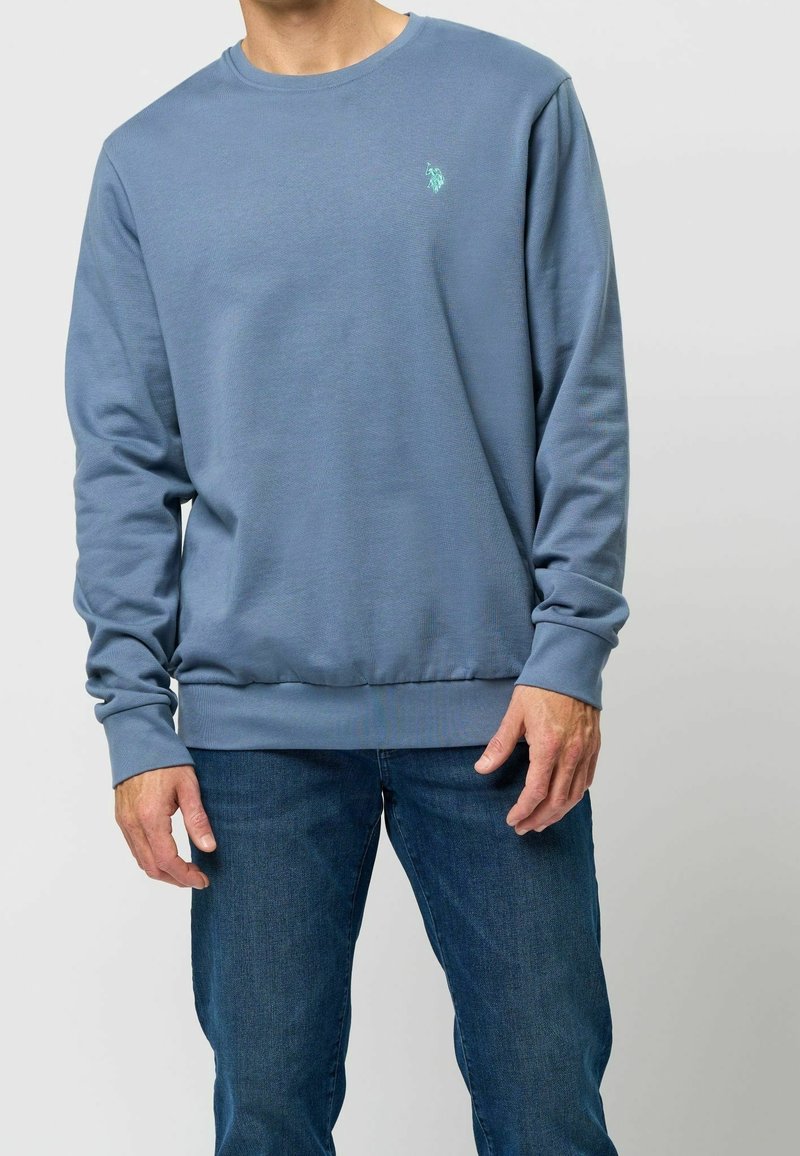 Homme portant un sweat-shirt bleu col rond avec un petit logo turquoise sur la poitrine et un jean bleu, debout devant un fond uni.