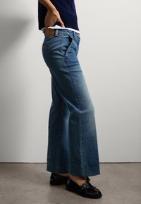 Jeans de denim azul de pierna ancha con cintura alta, que presentan costuras en contraste, un desgaste mínimo y una textura suave. Unos zapatos de cuero negros complementan el look.