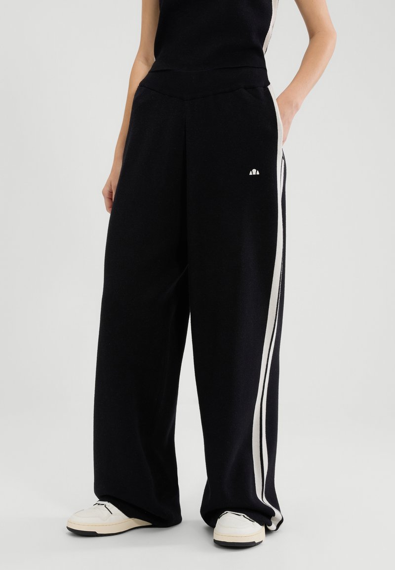 Schwarze, weit geschnittene Hose aus weichem Stoff, mit cremefarbenen Seitenstreifen und kleinem Logo auf der Vorderseite. Kombiniert mit weißen Turnschuhen.