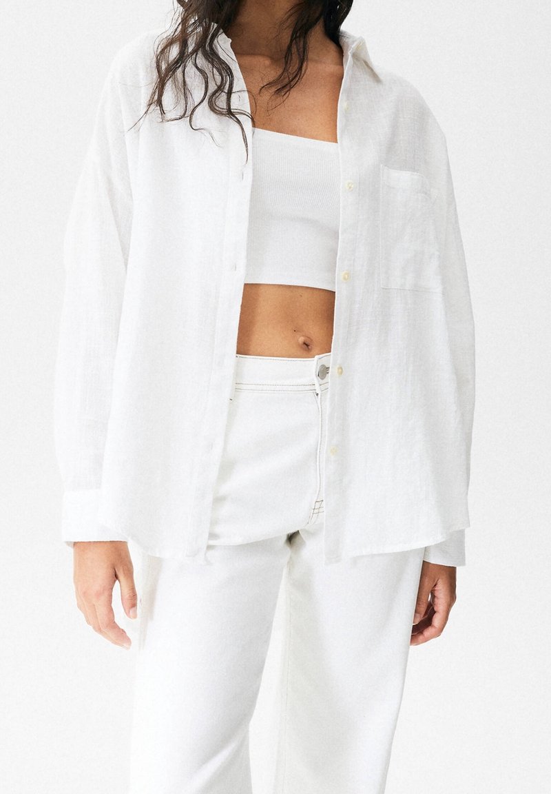 White linen button-up shirt with a relaxed fit, featuring long sleeves, a single chest pocket, worn over a white fitted tank top and pants.