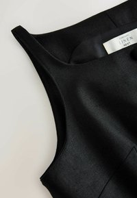 Haut sans manches en lin noir avec larges bretelles et une étiquette en tissu visible portant la mention "Contains Linen Next" sur un fond clair.