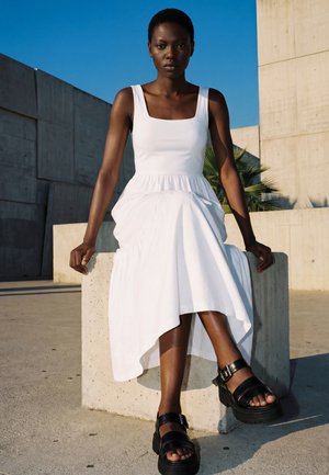 Woman with short hair wearing a sleeveless white dress and black platform sandals sits on a concrete block outdoors against blue sky.