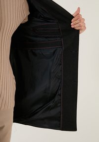 Interno di cappotto in lana nera con fodera liscia nera, cuciture rosse, una tasca e design strutturato. Mano che tiene il cappotto.