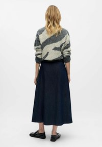 Kvinde med bølget blond hår iført en grå og hvid mønstret sweater, mørkeblåt midikjole og sorte loafers, stående mod en hvid baggrund.