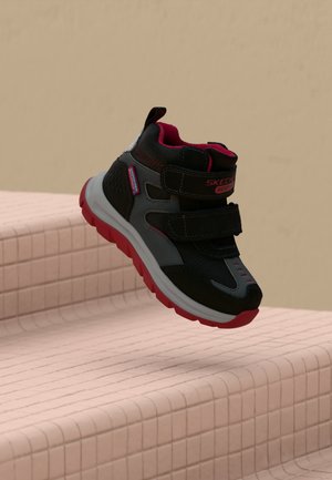 Chaussure pour enfant Skechers noire et grise avec des accents rouges et deux brides à scratch, représentée au-dessus de marches carrelées claires.