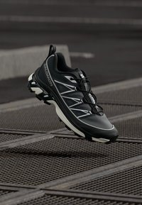 Sapato esportivo preto com parte superior texturizada, detalhes brancos, solado agressivo e sistema de atilhos seguro, projetado para desempenho e durabilidade.