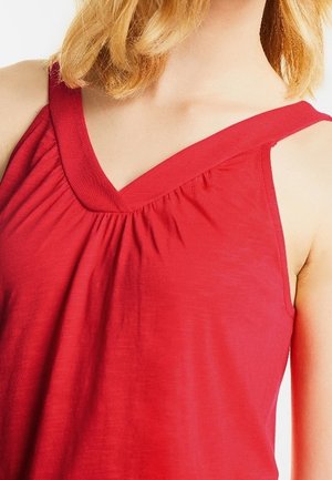 Kobieta w czerwonym topie bez rękawów, z dekoltem w kształcie litery V i marszczonym detalem pod kołnierzykiem.