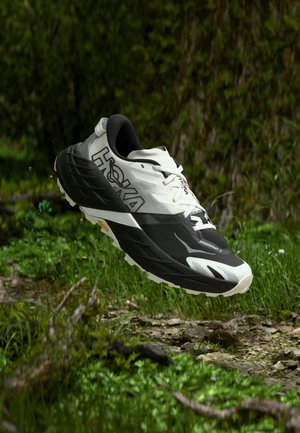 Juodos ir baltos spalvos Hoka Speedgoat 7 bėgimo takų bateliai skrenda virš žolėto, akmenuoto miško paviršiaus.