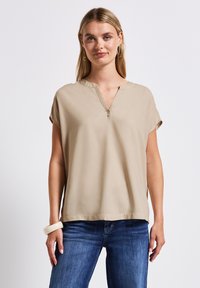 Beige Kurzarmoberteil mit V-Ausschnitt und lockerem Schnitt. Sanfte Textur und subtile seitliche Schlitze. Kombiniert mit blauen Jeans und einem klobigen Armband.