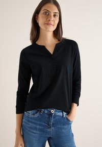 Musta pitkähihaiset paita, jossa on henley-kaarroke ja kolme nappia etupuolella. Pehmeä materiaali ja rento istuvuus, yhdistettynä sinisiin farkkuihin.