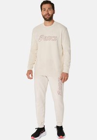 Ensemble sweat-shirt et pantalon couleur crème avec un logo "asics" rouge bien visible ; tissu doux avec des poignets et une taille côtelés ; chaussures de sport noires.