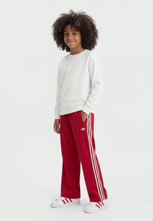Enfant aux cheveux bouclés, portant un sweat-shirt blanc, un pantalon de survêtement rouge Adidas avec des bandes blanches et des baskets blanches Adidas, debout et souriant.