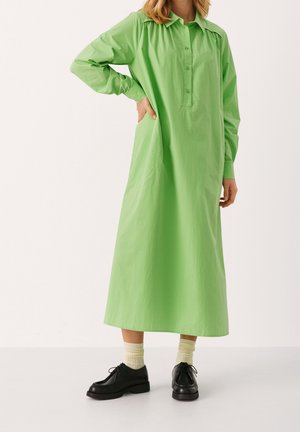 Femme portant une longue robe verte boutonnée, des chaussettes crème et des chaussures noires à lacets, debout avec une main sur la hanche.