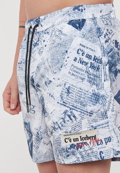 Costumi da bagno bianchi con motivo a stampa giornale blu, vita con cordoncino e un'etichetta con la scritta "C'è un Iceberg dal 1974."
