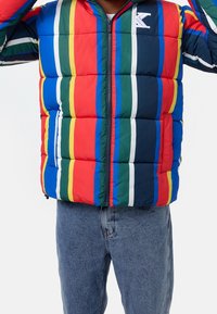 Giacca a vento imbottita con strisce multicolori in rosso, blu, verde e giallo. Presenta una chiusura con zip sul davanti e un dettagli logo sul petto.