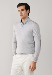 Homme portant un pull en V gris clair sur une chemise à col bleu clair et un pantalon blanc, debout devant un fond uni.