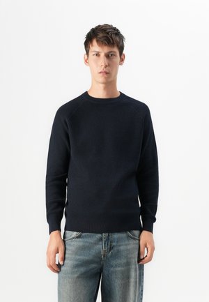 Jonge man in een gestructureerde donkerblauwe trui en vervaagde blauwe spijkerbroek, staande tegen een effen witte achtergrond.