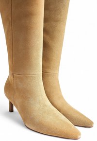 Botas hasta la rodilla en ante beige, con punta afilada y un tacón modesto. Textura suave con detalles de costura mínimos.