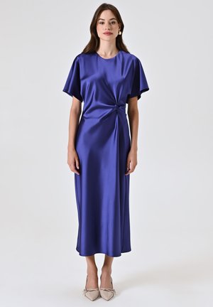 Abito lungo in satin viola con maniche corte, dettaglio a nodo torcigliato in vita e orlo dritto. Abbinato a tacchi beige.