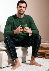 Zielona longsleeve z zapięciem na guziki; czarne i zielone spodnie piżamowe w kratę. Siedzi na kanapie, trzymając zielony kubek. Na boso.