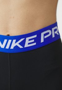 Crne atletske kratke hlače s plavim elastičnim pojasom na kojem stoji bijeli natpis "NIKE PRO". Prikaz u krupnom planu pokazuje glatku teksturu i usku prilagodbu.