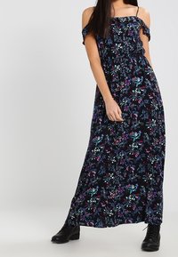 Robe maxi fleurie noire avec manches épaules dénudées, arborant un imprimé vibrant de fleurs bleues, violettes et blanches. Associée à des bottines noires.