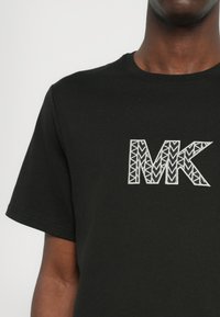 Čierne bavlnené tričko s veľkým vyšitým logom "MK" s geometrickými vzormi v sivej farbe. Klasický okrúhly výstrih a krátke rukávy.