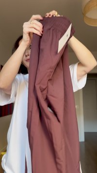 Person holding up maroon pants with elastic waistband and white tag, indoors in a softly lit room.