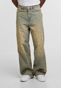 Muškarac u širokim izblijedjelim zelenim trapericama s crnim mrljama od boje i bijelim tenisicama, stoji ispred obične pozadine.