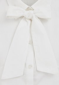 Bijela bluza s mašnom na ovratniku, izrađena od glatke tkanine. Ima kopčanje na gumbe i uredno posložene vrhove ovratnika.