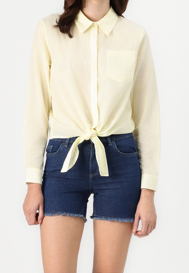 Chemise jaune boutonnée à manches longues, nouée à la taille. Associée à un short en denim foncé avec ourlet effiloché et poches à l'avant.