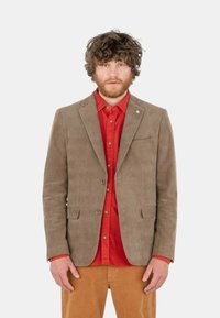 Blazer en velours côtelé marron avec revers crantés, fermeture à un bouton, et deux poches avant, porté sur une chemise rouge et un pantalon beige.