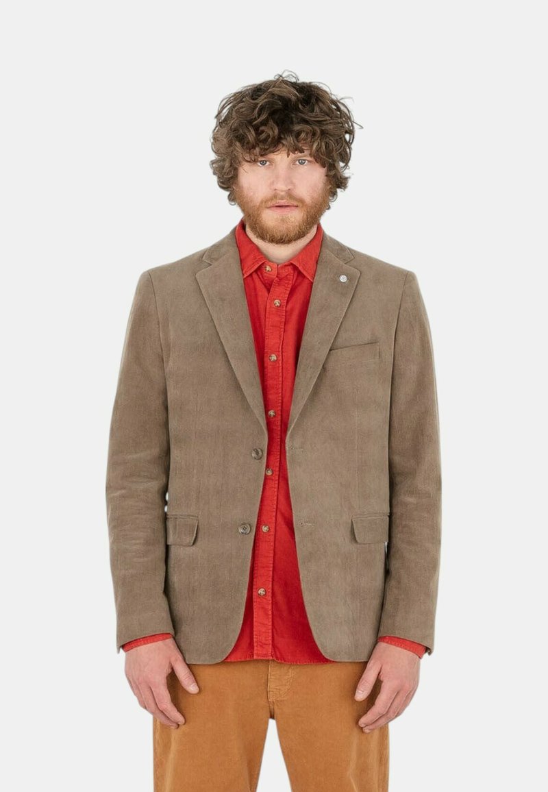Blazer en velours côtelé marron avec revers crantés, fermeture à un bouton, et deux poches avant, porté sur une chemise rouge et un pantalon beige.